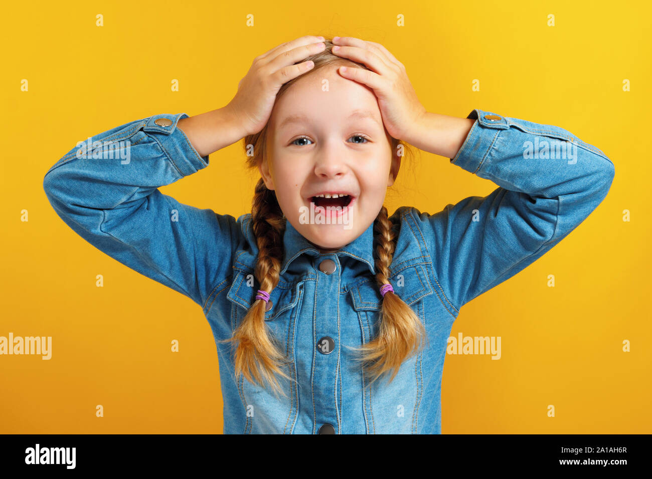 Little girl holds hands behind her head on a yellow background. The child is amazed, surprised, emotional. Stock Photo