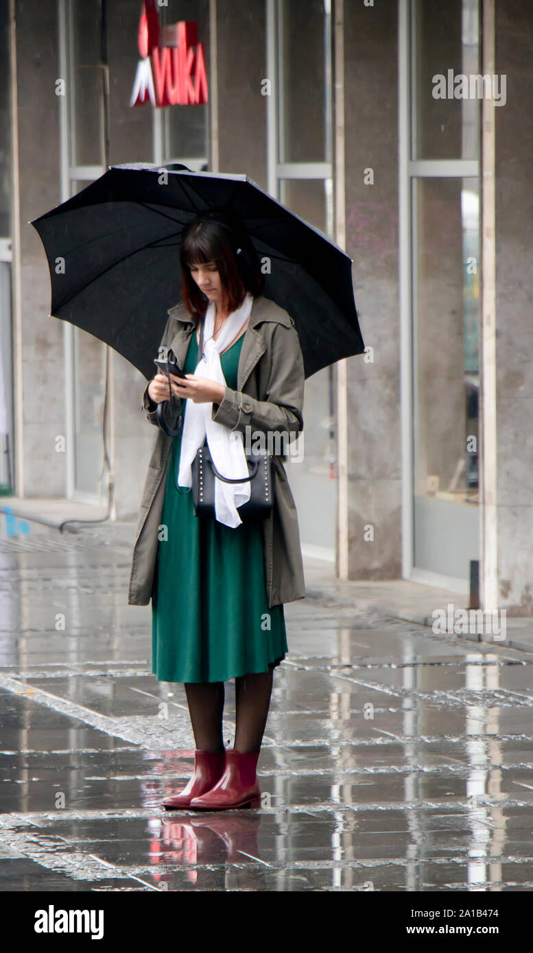 Belgrade, Serbia- September 24, 2018: Young elegant woman standing under umbrella on a rainy city street and looking at her mobile phone Stock Photo