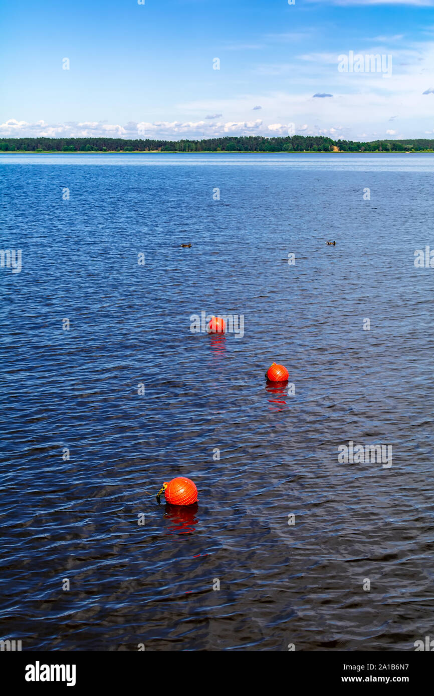 Three bright orange buoys floating on river water surface with woods at the horizon. Set of three orange safety buoys stretching into the distance. Stock Photo