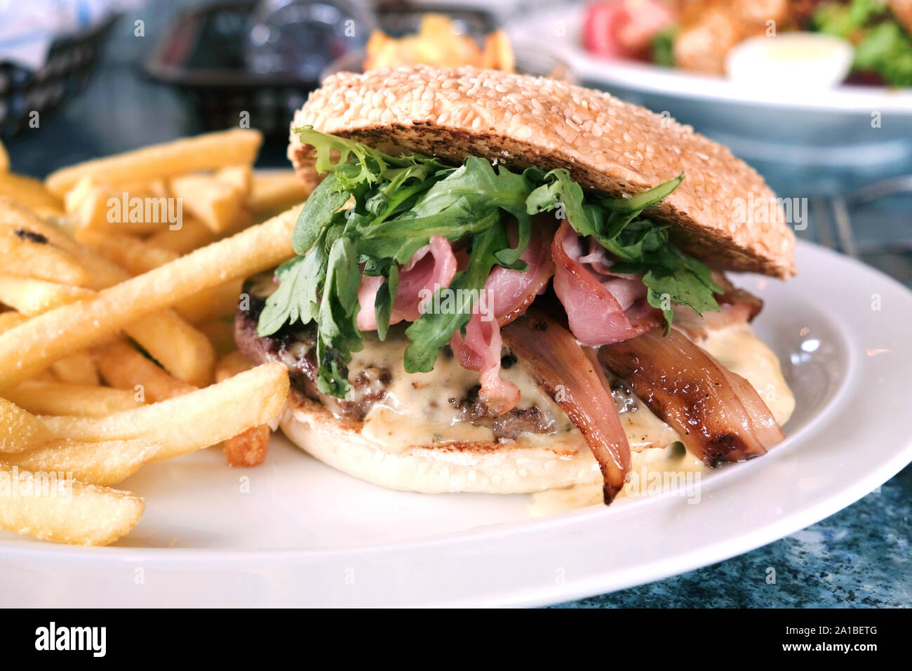 A tasty looking gourmet beefburger and french fries or chips served up on a restaurant table. It has green leaf and a grilled bacon topping Stock Photo