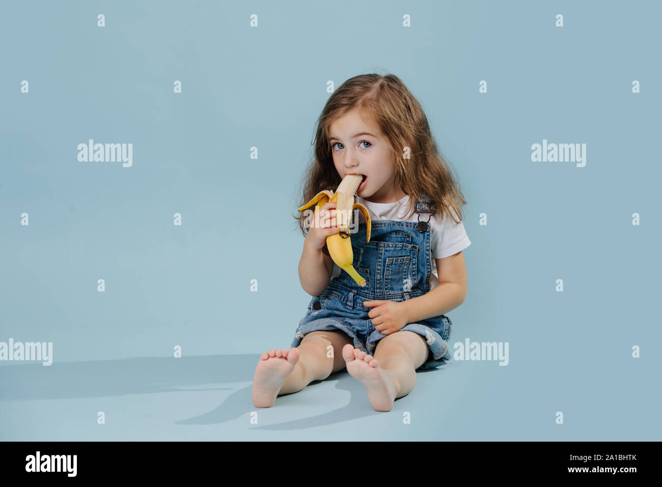 Little girl is eating banana, while sitting on the floor over blue background Stock Photo