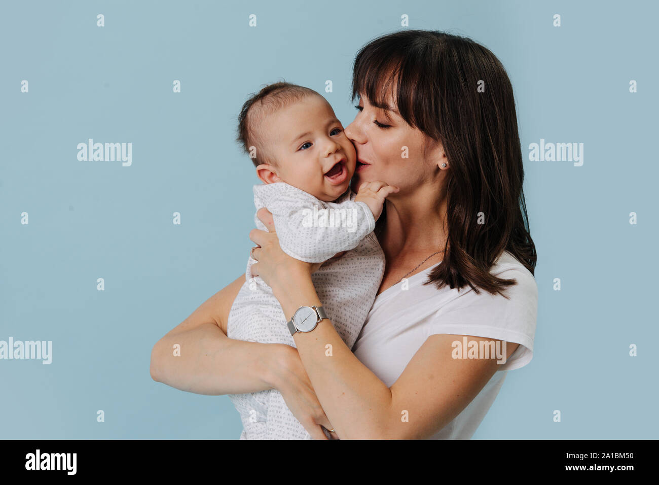 Mother is holding on hands, kissing her small jolly infant baby over blue Stock Photo