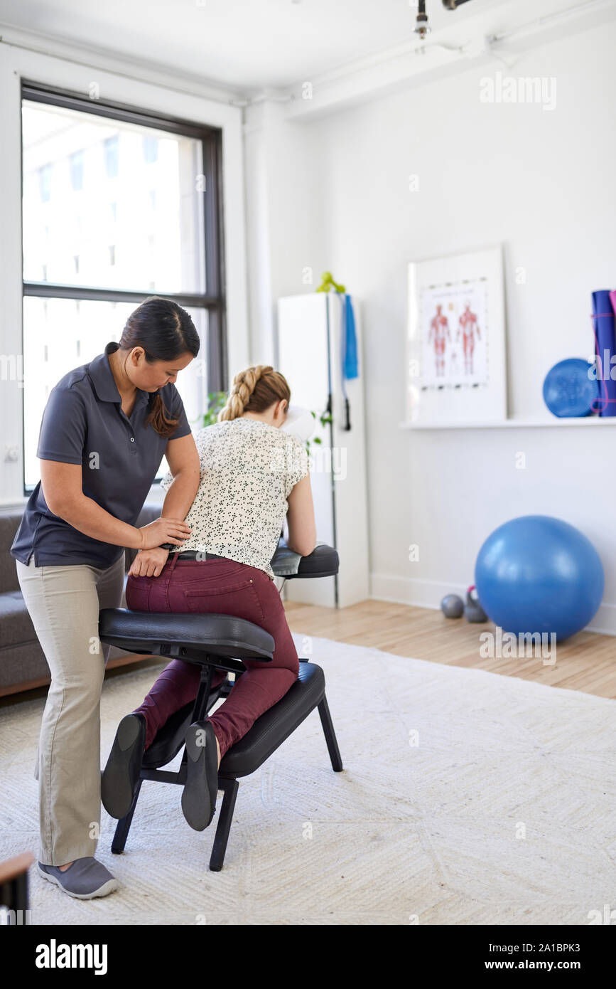 Chinese woman massage therapist giving a neck and back pressure treatment to an attractive blond client at her workplace in a bright office Stock Photo