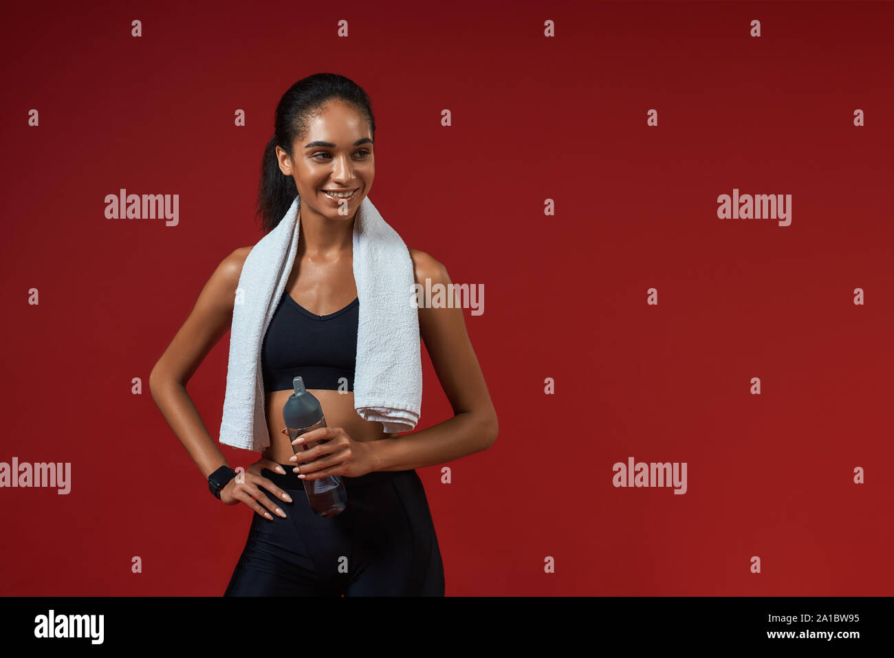 Cheerful afro american woman in sports clothing with towel on her shoulders holding bottle of water and smiling while standing against red background. Sport concept. Active lifestyle Stock Photo