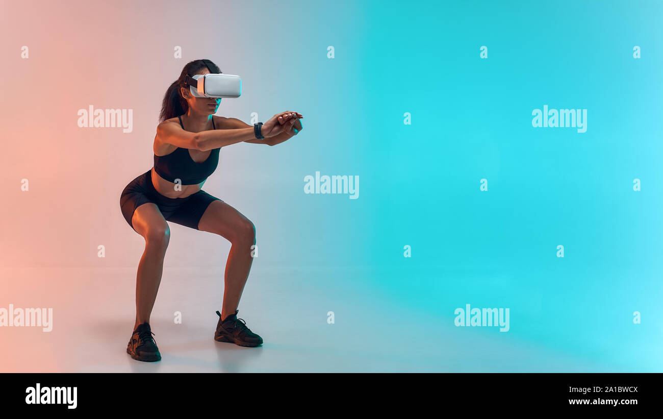 Full length of young sporty woman in sports clothing doing squat while wearing virtual reality glasses. Studio shot. Future technology Stock Photo
