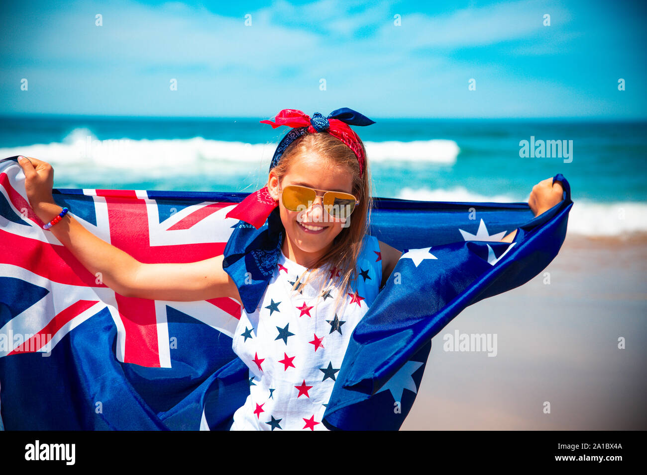 Happy Australian girl carries fluttering blue white red Australian flag against blue sky and ocean background Stock Photo
