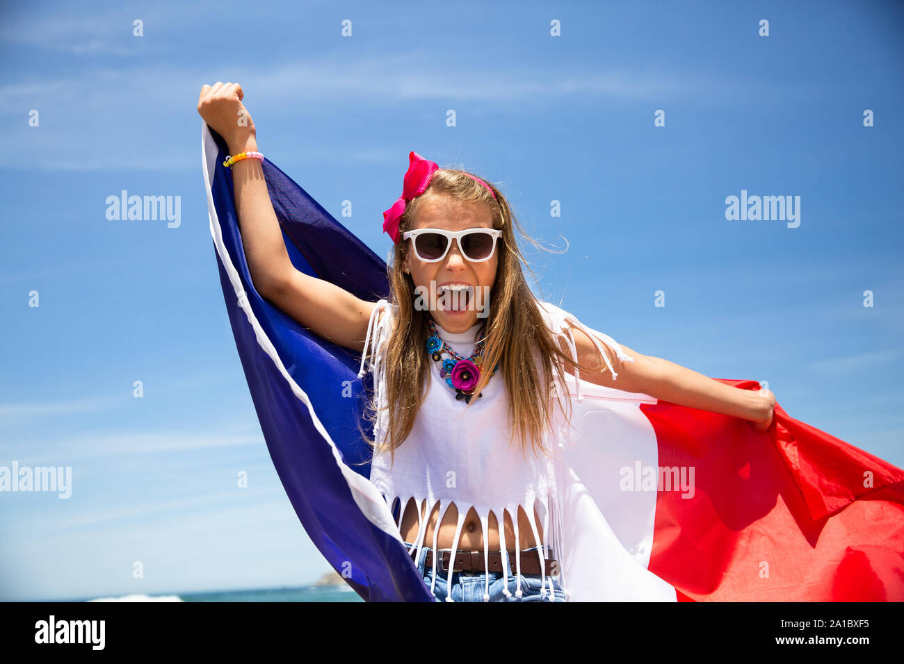 Happy French girl carries fluttering blue white red French flag of France against blue sky and Atlantic ocean background Stock Photo