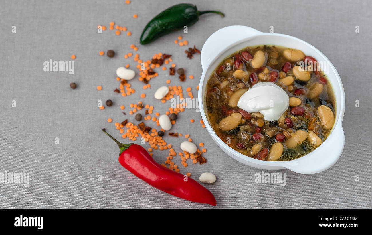 Mexican soup of seven kinds of beans with sour cream, close-up, on a gray linen background surrounded by red pepper and beans Stock Photo