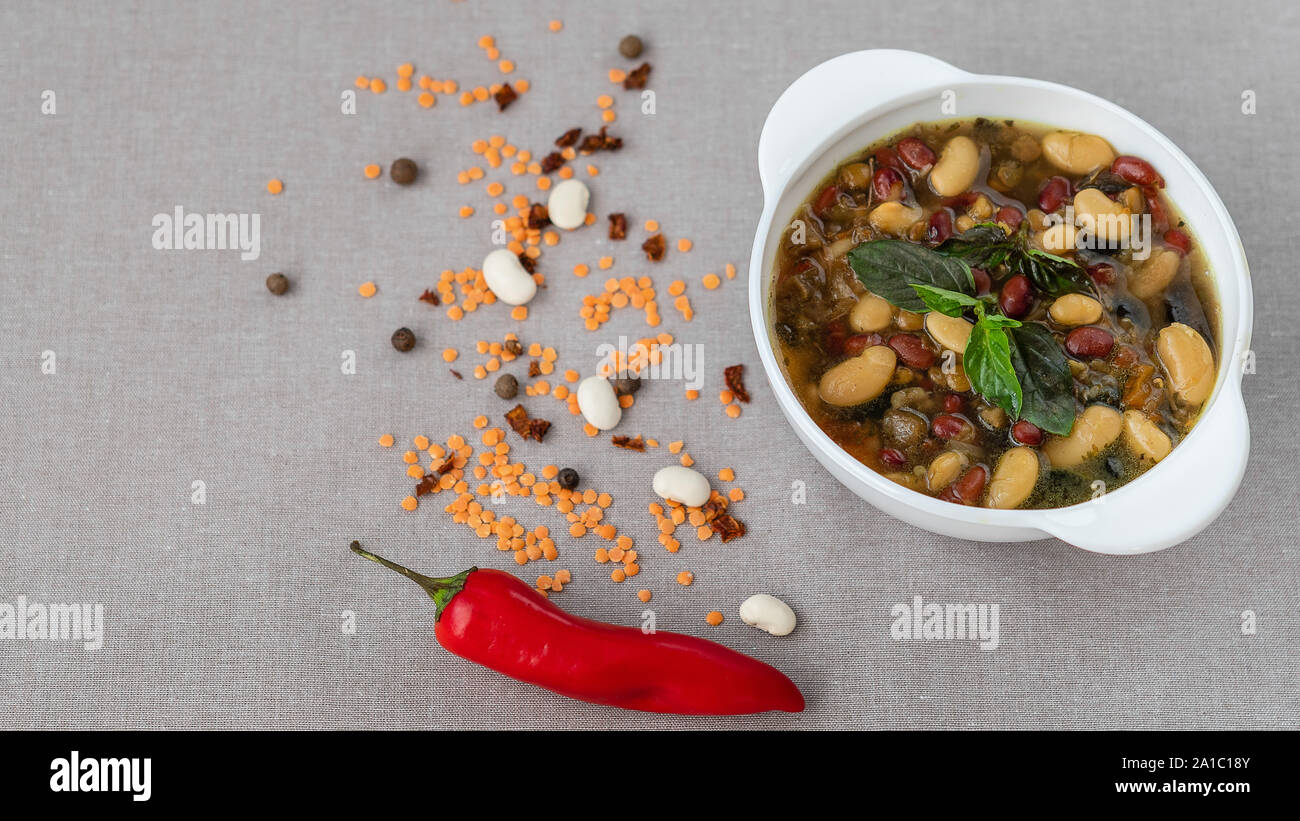 Mexican soup of seven kinds of beans with basil, close-up, on a gray linen background surrounded by red pepper and beans Stock Photo