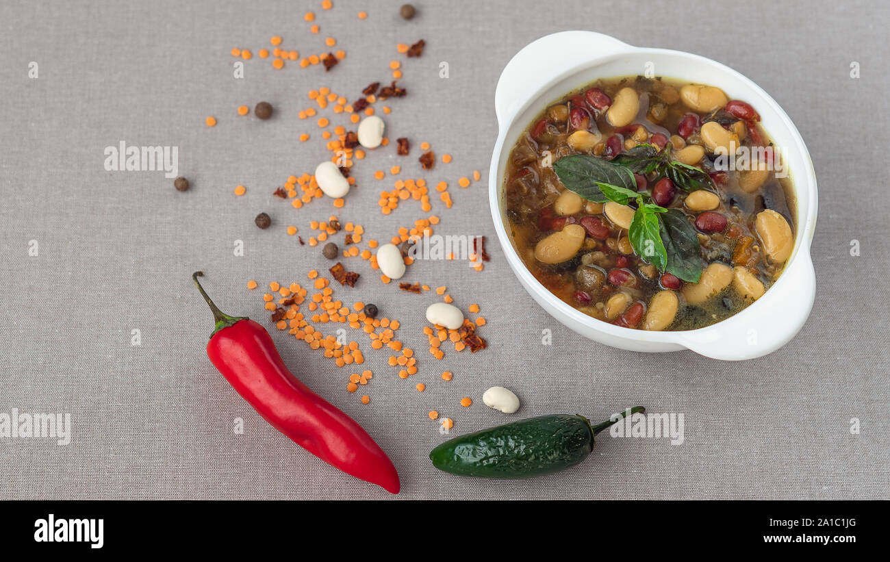 Mexican soup of seven kinds of beans, close-up, on a gray linen background surrounded by red and green peppers and beans Stock Photo