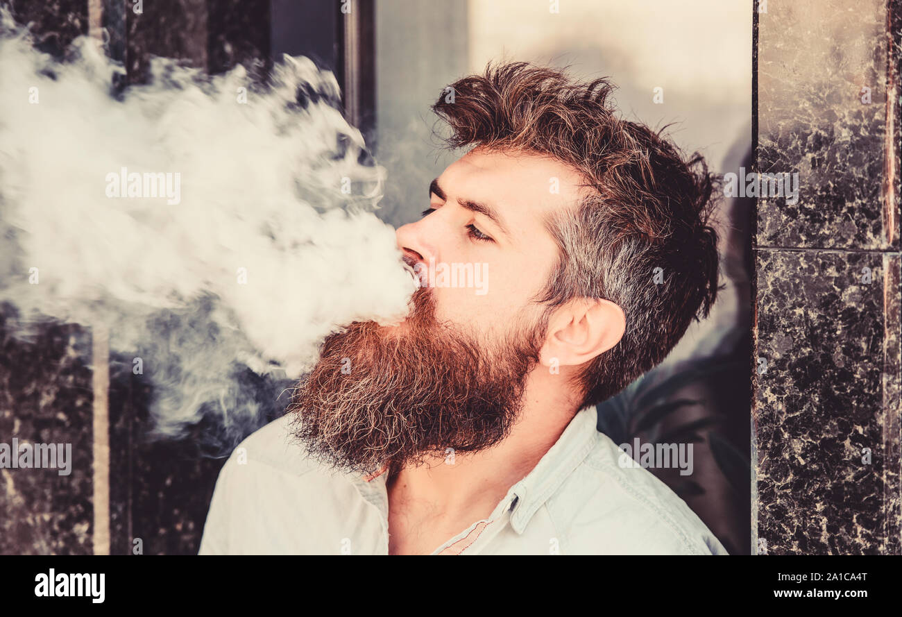 Smoking electronic cigarette. Stress relief concept. Smoking device. Man long beard relaxed with smoking habit. Clouds of flavored smoke. Bearded man smoking vape. Man with beard breathe out smoke. Stock Photo