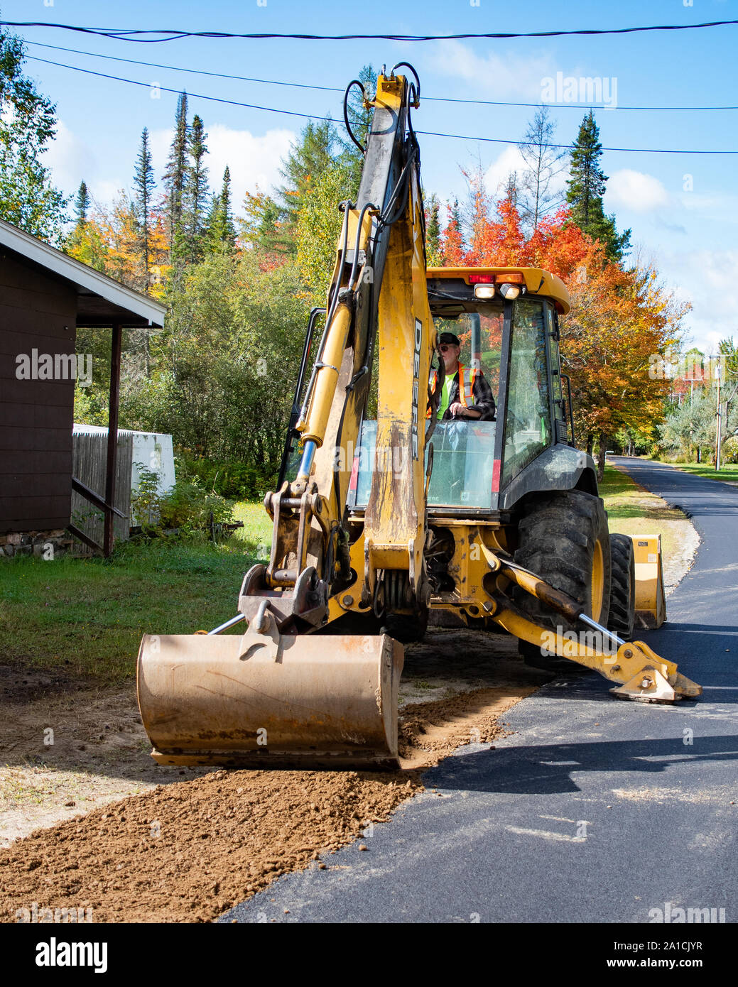 A wheeled backhoe excavator working on smoothing the shoulder and edges of a village street in Speculator, NY USA Stock Photo