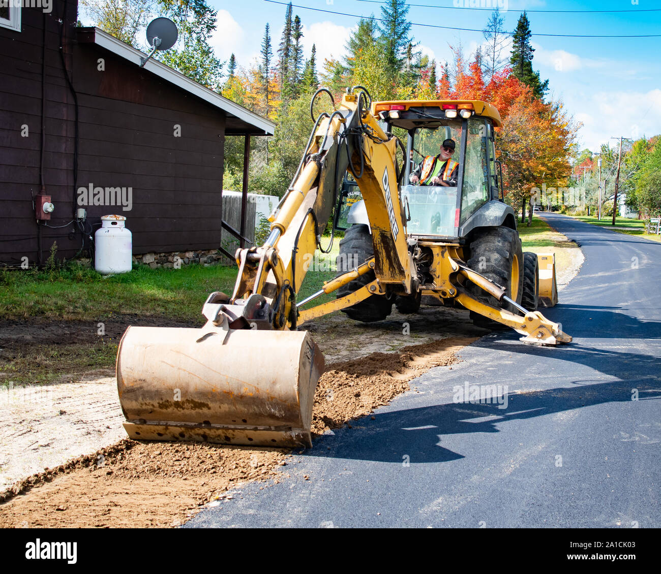 A wheeled backhoe excavator working on smoothing the shoulder and edges of a village street in Speculator, NY USA Stock Photo