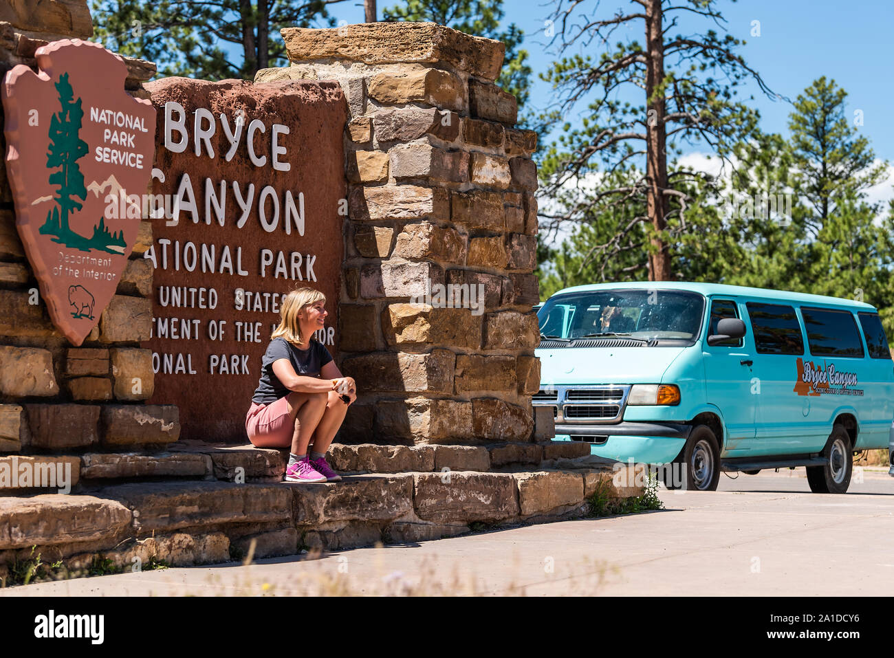 Bryce Canyon City, USA - August 1, 2019: Tourists taking pictures by national park entrance sign with woman posing for photo and tour bus Stock Photo
