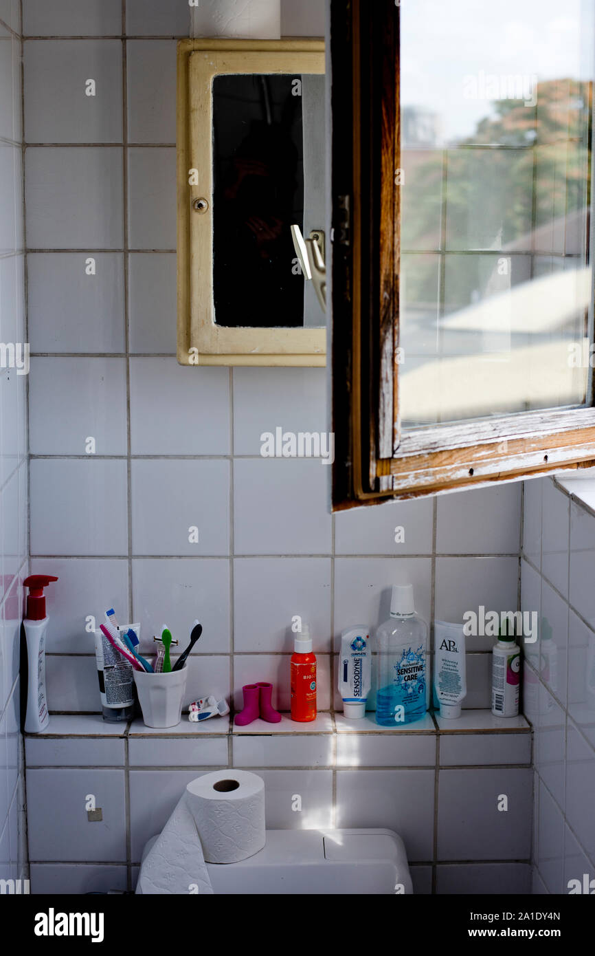 A typical, white-tiled bathroom in Antwerp, Belgium, with a mirror hanging above the toiletries and an open window reflecting rooftops and trees. Stock Photo