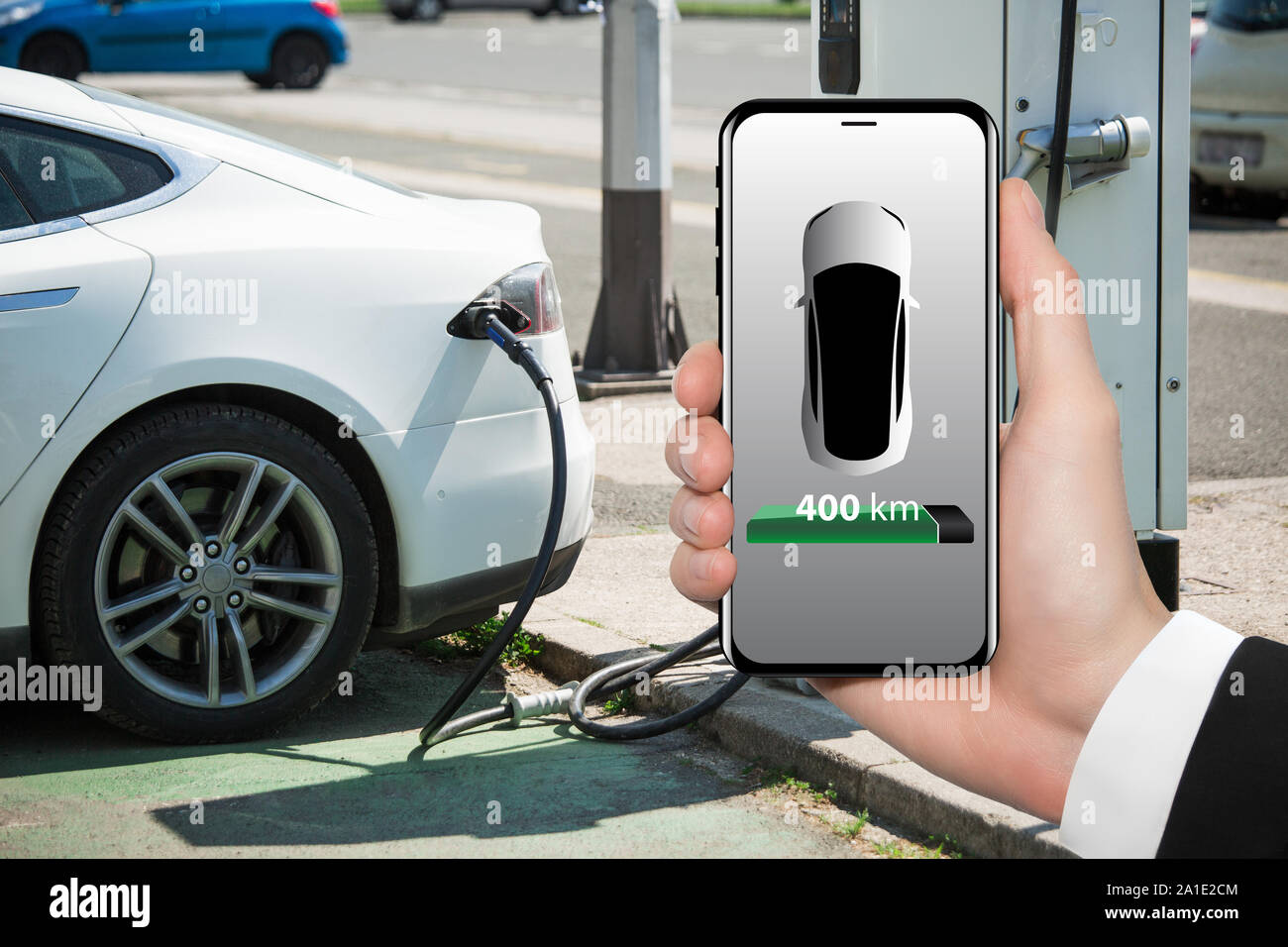 Hand with phone on a background of electric car charging point. On a device screen indicator of power reserve. Stock Photo