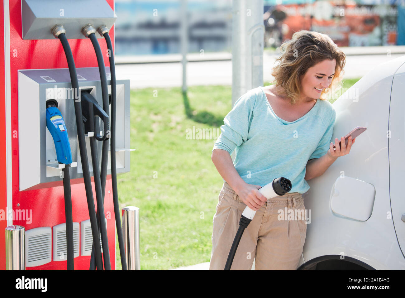 Young woman is standing near the electric car. The car is charging at the charging station for electric vehicles. Stock Photo
