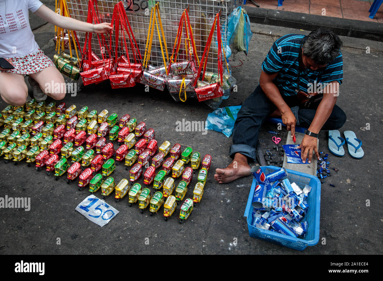 BANGKOK, THAILAND: A street vendor sits on the ground as he makes Tuk Tuk models out of soft drink cans on Khao San Road in Bangkok, Thailand on August 22nd, 2019. Bangkok's bustling Khao San Road - a strip famous among tourists for its budget hostels, street food and market stalls - is set for a £1.28M face lift in October this year. For many backpackers Khao San is the starting point for their travels across Southeast Asia. It is a place to meet fellow travellers, dine out on pad thai served from street carts, sip cocktails from small brightly-coloured buckets or perhaps get an ill-advised Stock Photo