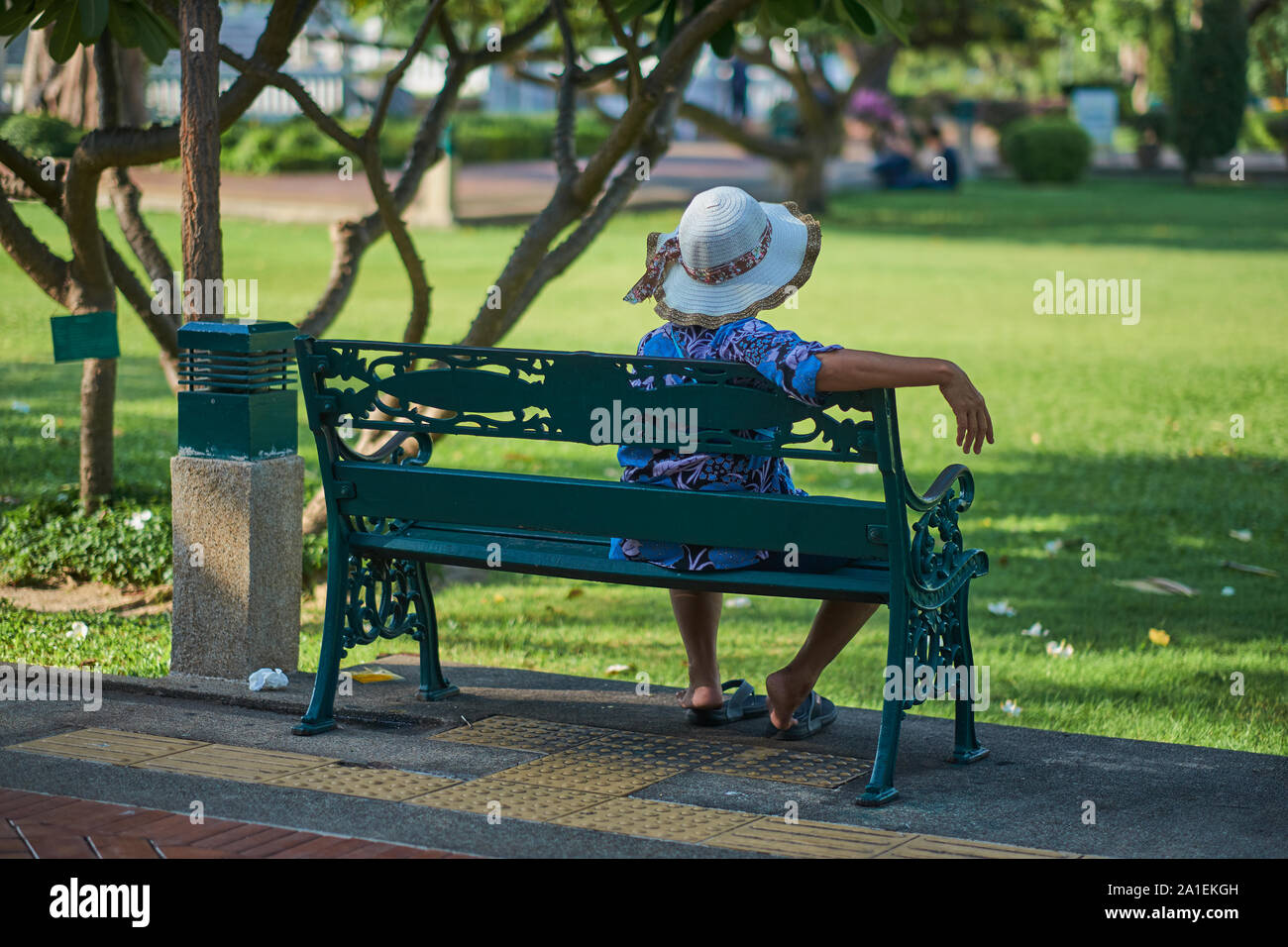 A Thai woman wearing a hat relaxes on a bench in Santichaiprakarn Park, in Banglamphoo area, Bangkok, Thailand Stock Photo