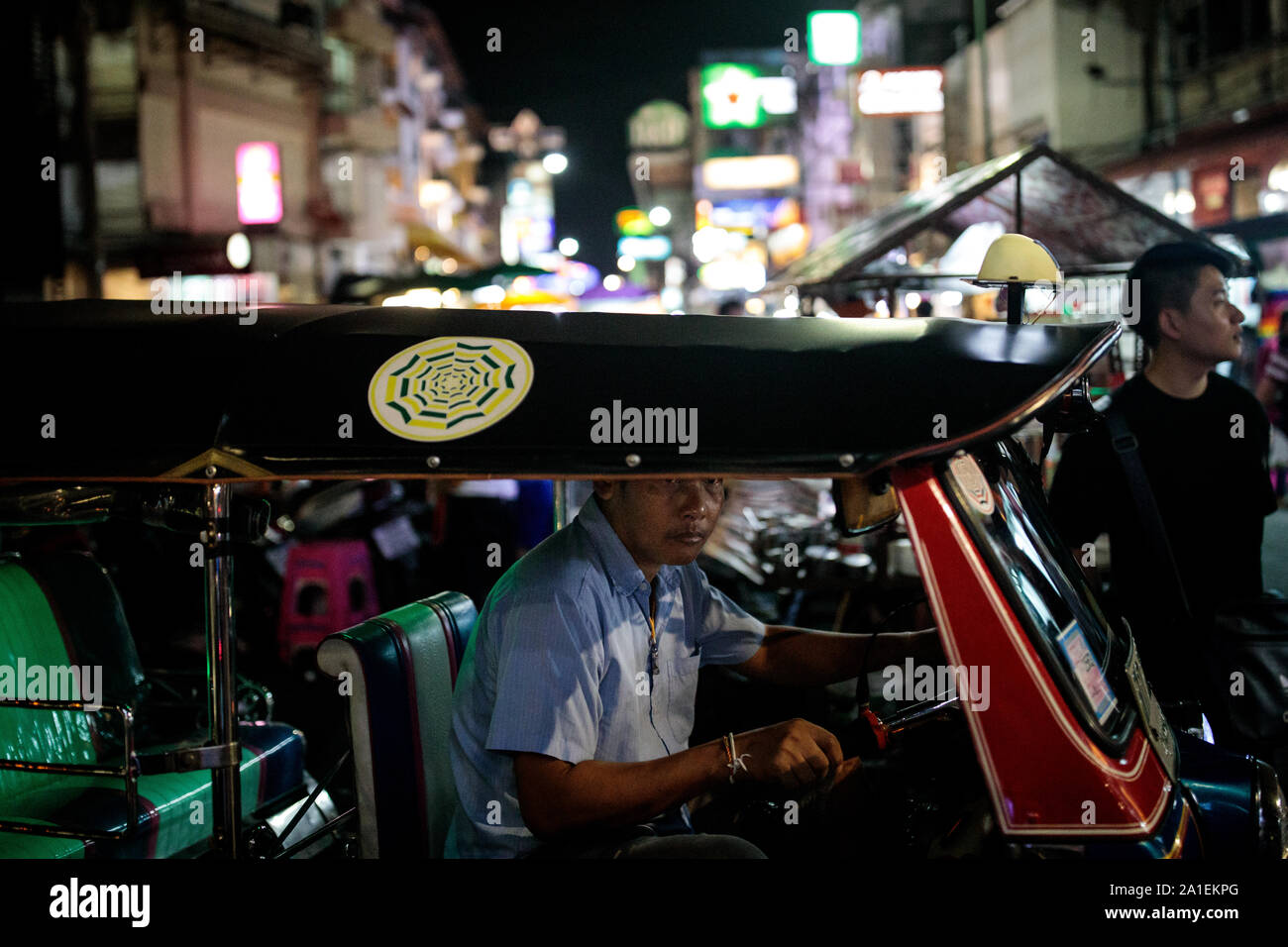 BANGKOK, THAILAND: A tuk tuk driver sits in his vehicle at the end of Khao San Road in Bangkok, Thailand on August 27th, 2019. Bangkok's bustling Khao San Road - a strip famous among tourists for its budget hostels, street food and market stalls - is set for a £1.28M face lift in October this year.   For many backpackers Khao San is the starting point for their travels across Southeast Asia. It is a place to meet fellow travellers, dine out on pad thai served from street carts, sip cocktails from small brightly-coloured buckets or perhaps get an ill-advised tattoo.   A place both garish and ch Stock Photo