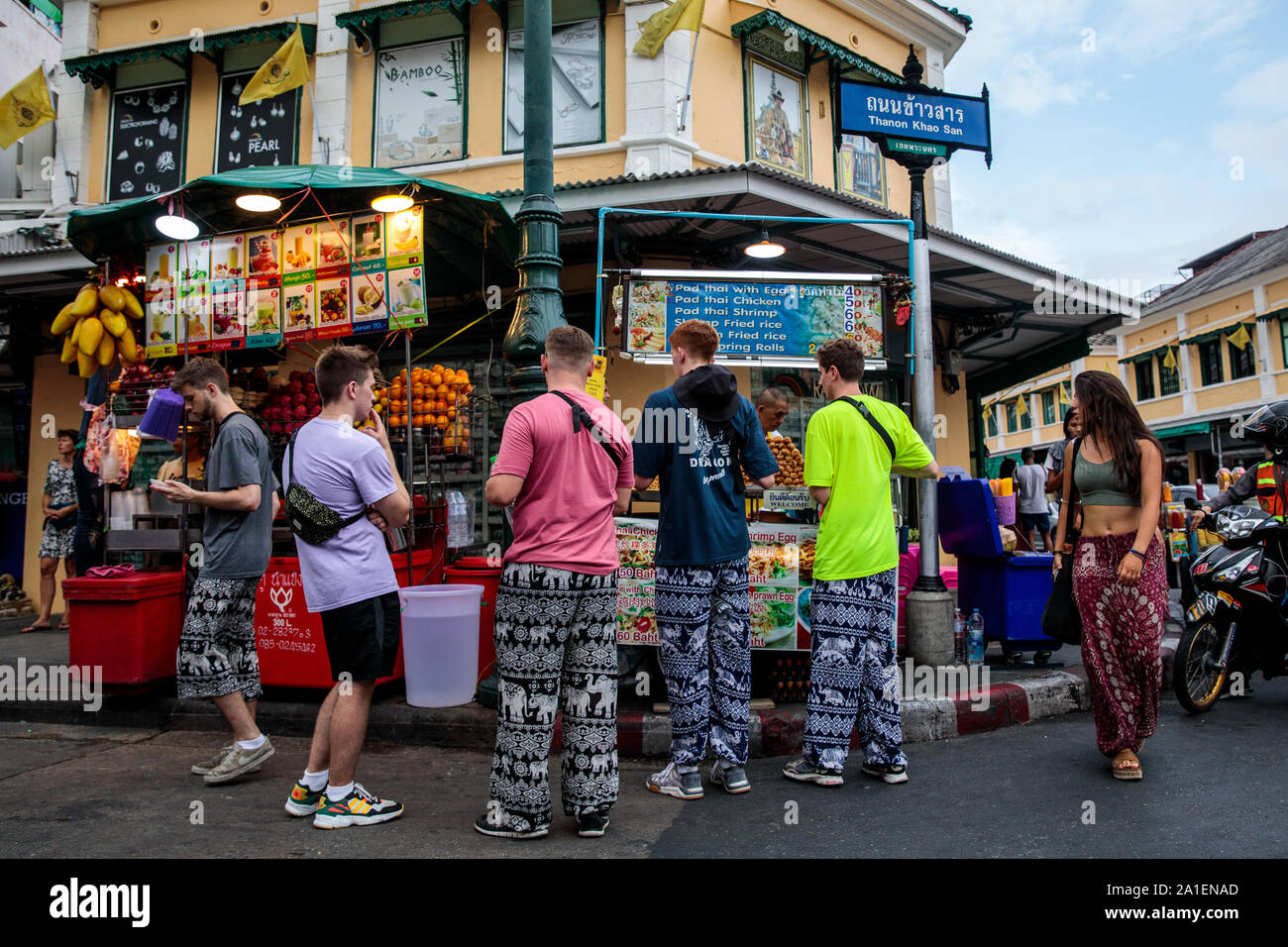 BANGKOK, THAILAND: Tourists wearing elephant-adorned trousers grab food at a stall on Khao San Road in Bangkok, Thailand on August 22nd, 2019. Bangkok's bustling Khao San Road - a strip famous among tourists for its budget hostels, street food and market stalls - is set for a £1.28M face lift in October this year.   For many backpackers Khao San is the starting point for their travels across Southeast Asia. It is a place to meet fellow travellers, dine out on pad thai served from street carts, sip cocktails from small brightly-coloured buckets or perhaps get an ill-advised tattoo.   A place bo Stock Photo
