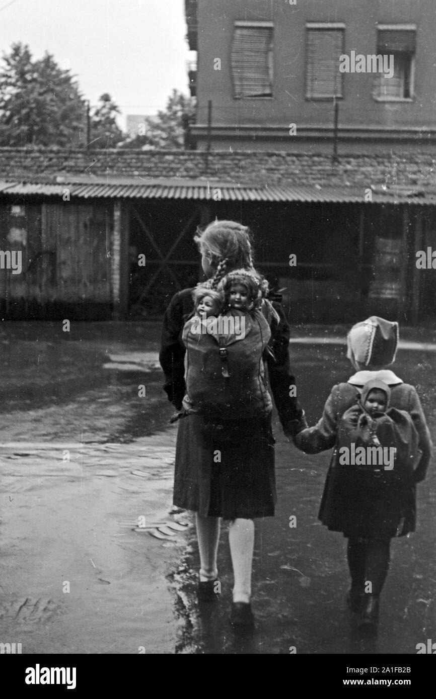 Zwei Mädchen mit ihren Puppen im Gepäck auf dem Weg durch das verregnete Berlin, Deutschland 1949. Two little girls with their dolls in a backpack on their way throuhgh the rainy Berlin, Germany 1949. Stock Photo