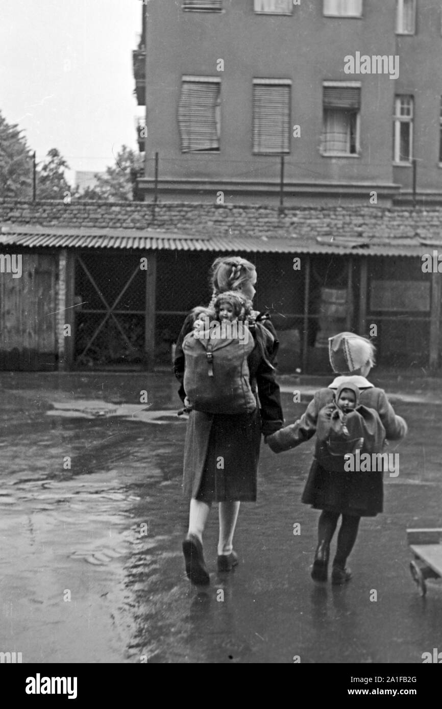 Zwei Mädchen mit ihren Puppen im Gepäck auf dem Weg durch das verregnete Berlin, Deutschland 1949. Two little girls with their dolls in a backpack on their way throuhgh the rainy Berlin, Germany 1949. Stock Photo