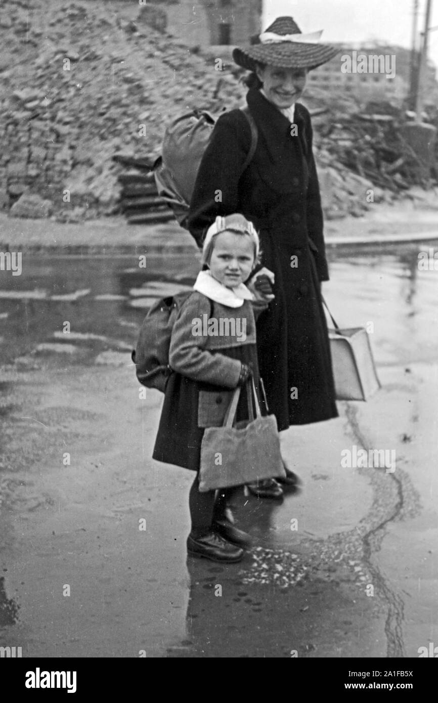 Mädchen mit ihrer Puppe im Gepäck und ihrer Mutter auf dem Weg durch das verregnete Berlin, Deutschland 1949. Little girl with her dolls in a backpack and her mother on her way throuhgh the rainy Berlin, Germany 1949. Stock Photo