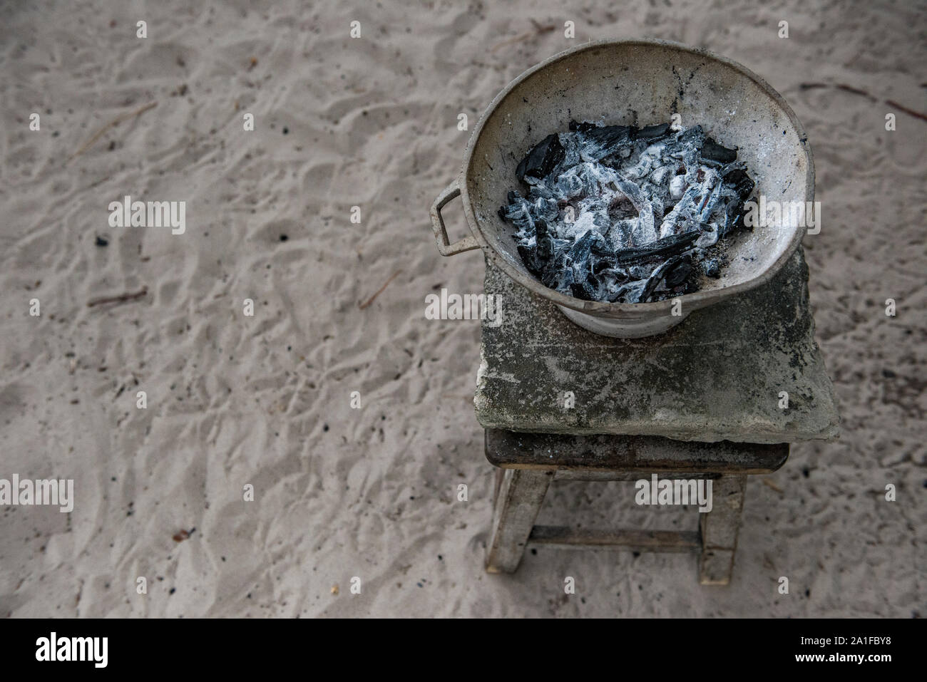 Old pan filled with burnt charcoal over wood bench on sandy floor Stock Photo