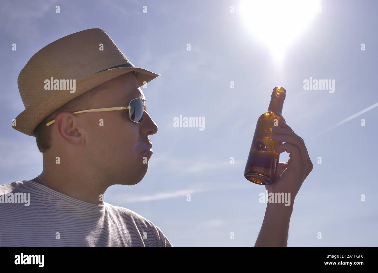 Attractive young man in a hat examines a brown bottle with a drink, outdoors against a blue sky Stock Photo