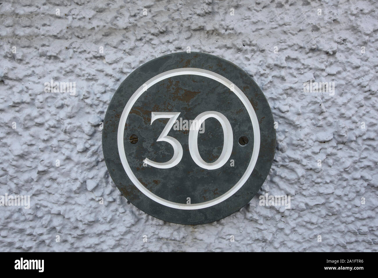 Building number thirty (30) on a round slate plaque set against a grey pebble dashed wall Stock Photo