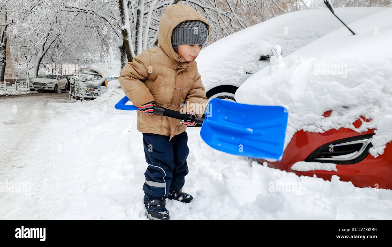 Close up shot of smiling charming boy in beige jacket and grey hat helping to clean up the snow covered red car after snow storm using big blue shovel Stock Photo