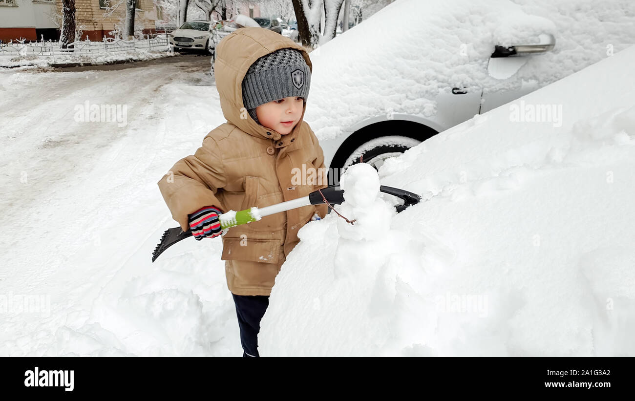 Close up photo of cute smiling boy in warm gloves hat and jacket helping to clean up snow covered parents red car after blizzard using big brush . In Stock Photo