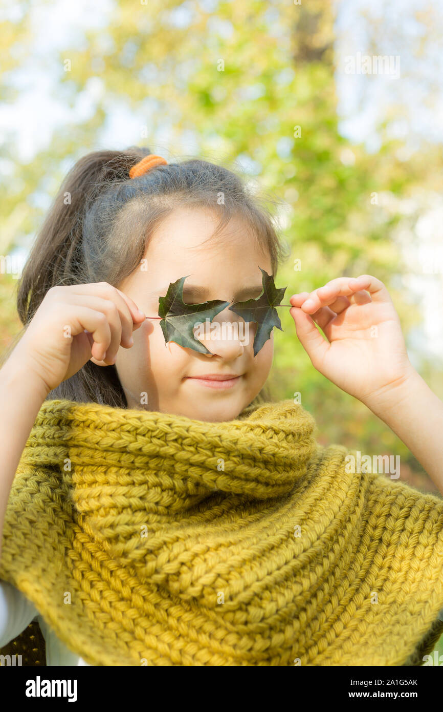 Smiling little girl covering eyes with green leaves. Kid autumn