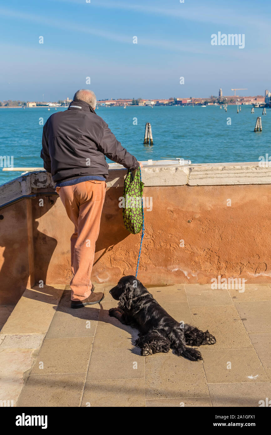 VENICE, ITALY - NOVEMBER 01, 2014: A man with a black dog on the waterfront looks into the distance across the Strait to the island Stock Photo