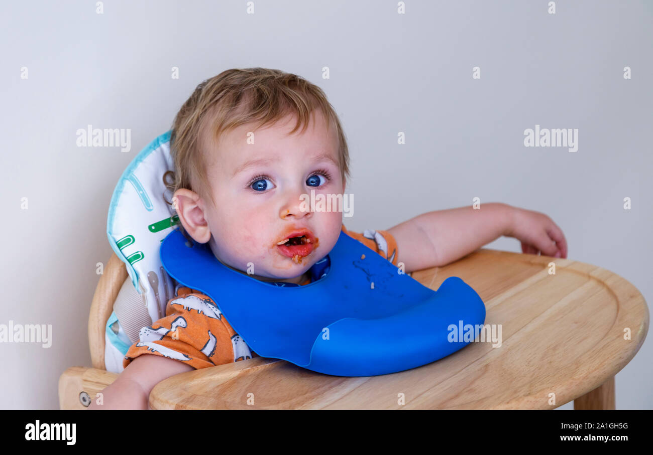 A 1 year old Caucasion blonde baby boy with a messy face makes a mess being fed in his high chair with a blue plastic pelican bib as he learns to eat Stock Photo