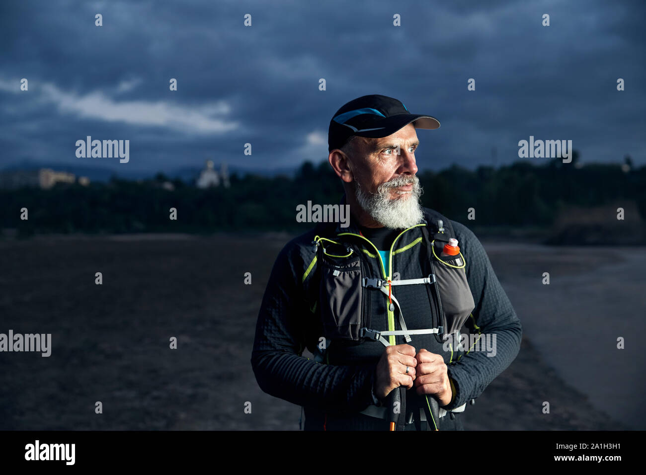 Portrait of elderly athlete man with grey beard and trekking poles at dark cloudy sky background Stock Photo