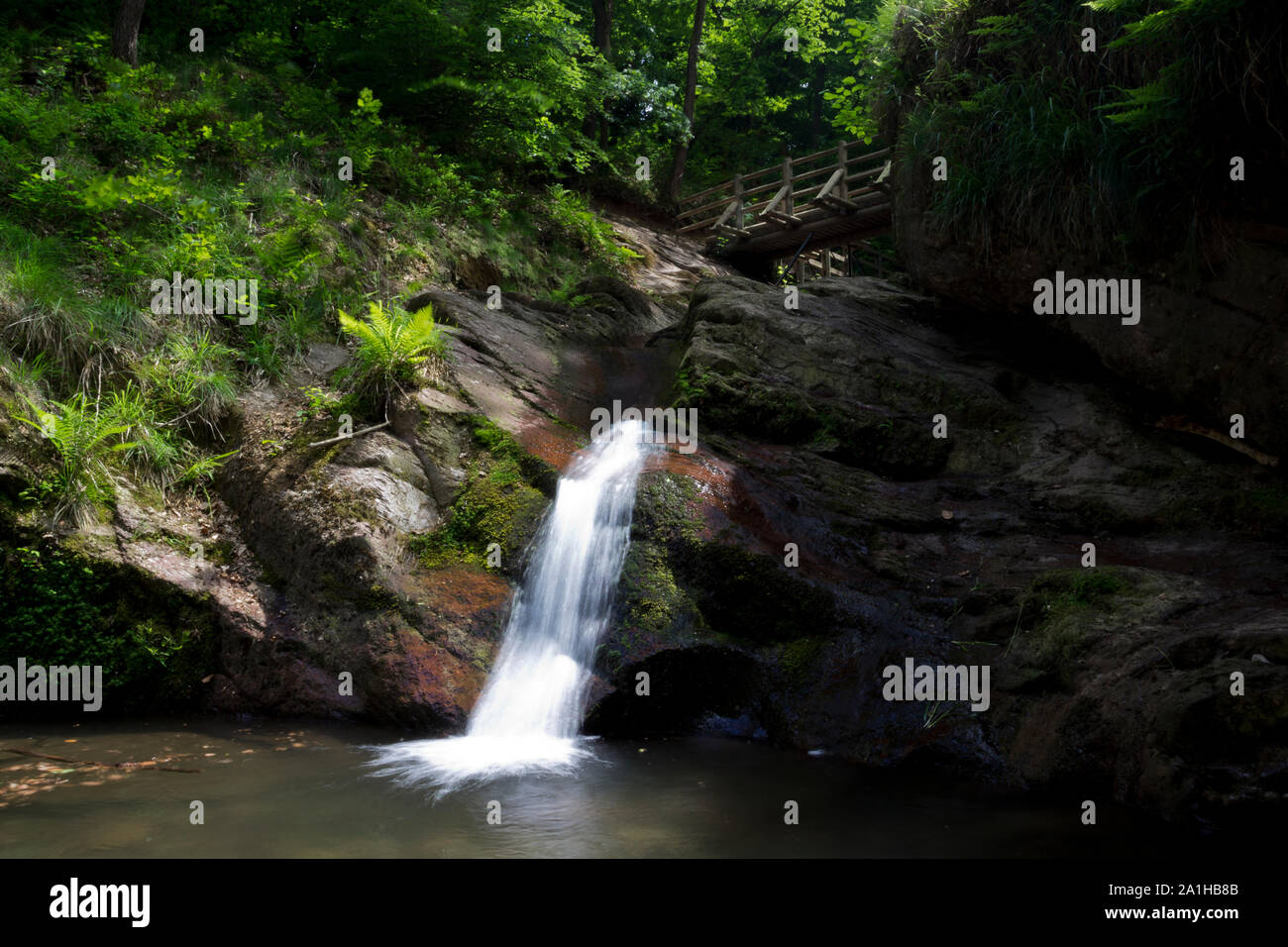 Waterfall passing under a wooden walkway bridge looking like a veil because of long exposure and stacked ND and polarizer filter surrounded by green. Stock Photo