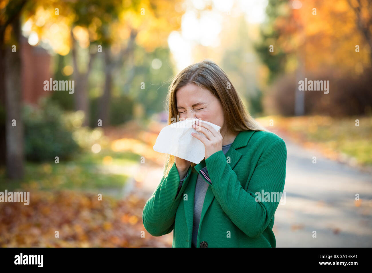 Sick young woman with cold and flu standing outdoors, sneezing, wiping nose with handkerchief, coughing. Autumn street background Stock Photo