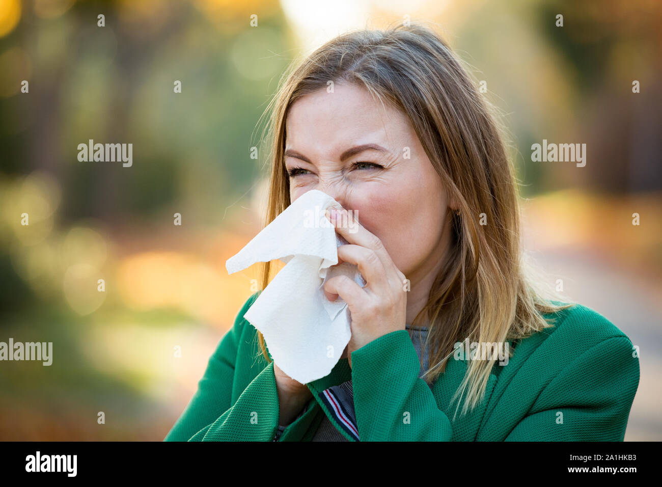 Sick young woman with cold and flu standing outdoors, sneezing, wiping nose with handkerchief, coughing. Autumn street background Stock Photo
