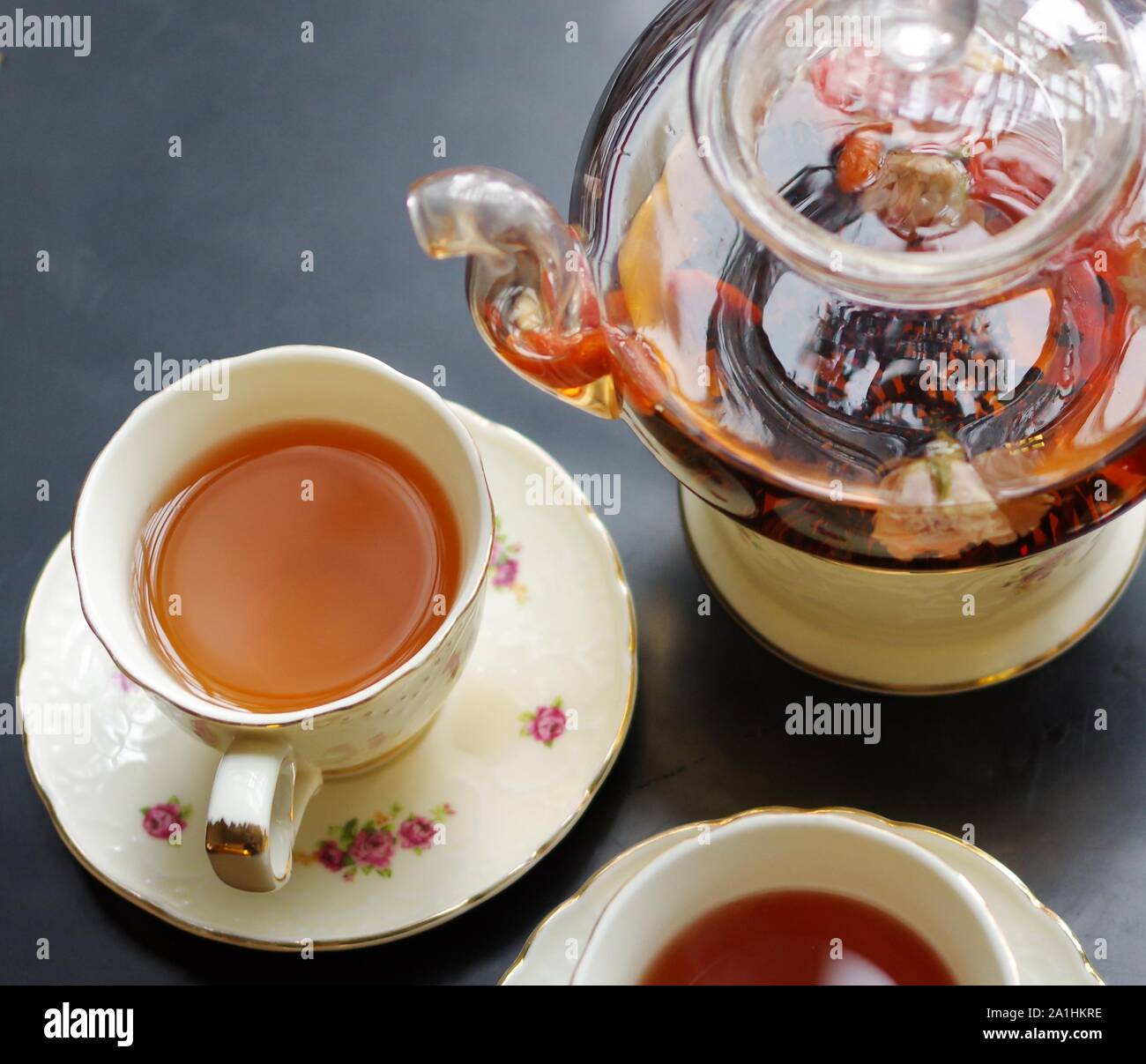 Black tea with goji (chinese wolfberries) and jasmine in glass teapot and porcelain cups with flower pattern saucers on black table Stock Photo