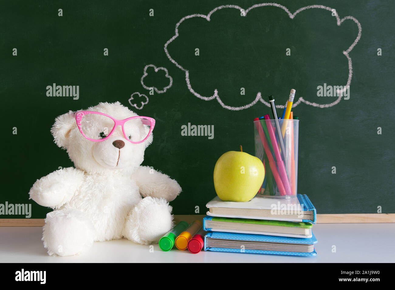 Cute teddy bear wearing glasses next to a pile of books and an apple in a classroom. Drawn thought bubble on a blackboard with empty copy space Stock Photo