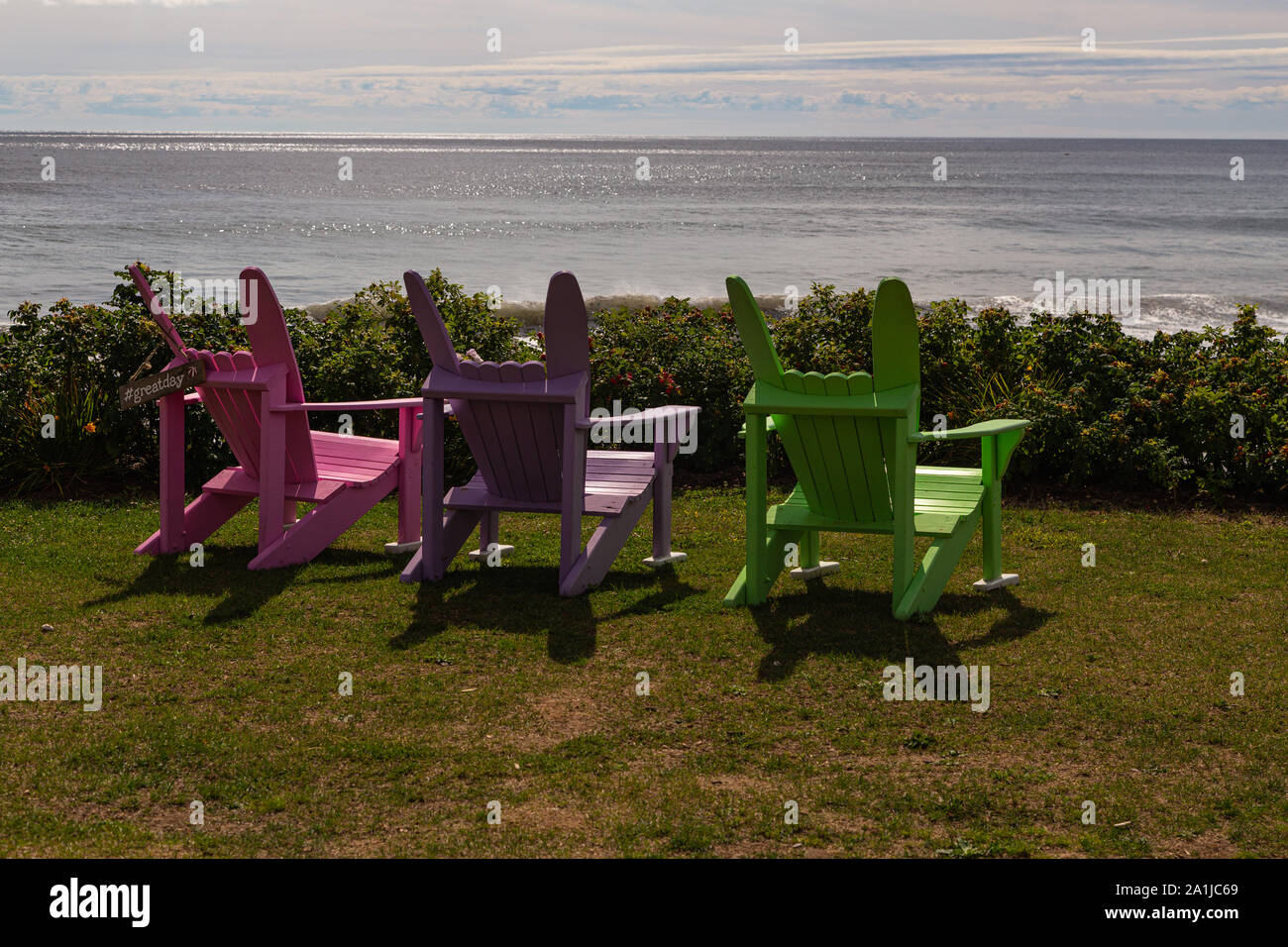 Three empty, brightly coloured lawn chairs on the grass, overlooking the Atlantic Ocean at White Point Beach, Nova Scotia, Canada. Stock Photo