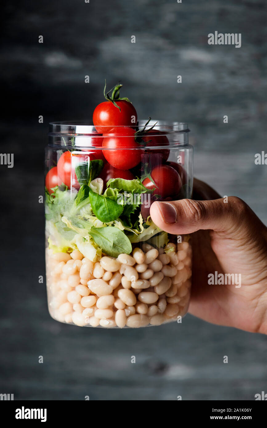 closeup of a young caucasian man with a jar salad in his hand, made with cherry tomatoes, lettuce and cooked white beans, against a gray background Stock Photo