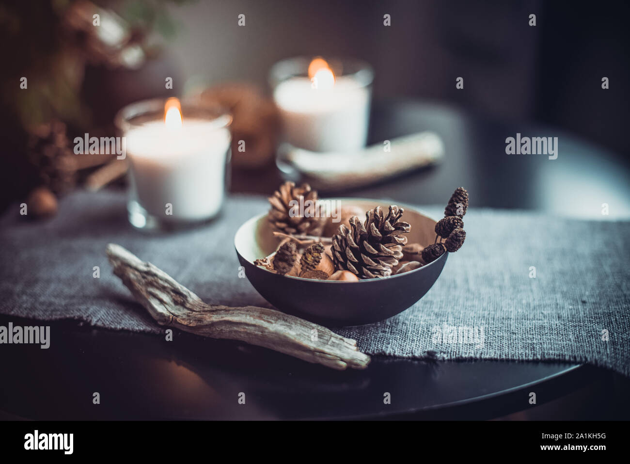 Burning candles and natural decor of cones, nuts in black bowl on the gray fabric napkin on the black table. Cozy atmosphere at home. Kinfolk Hygge Sl Stock Photo
