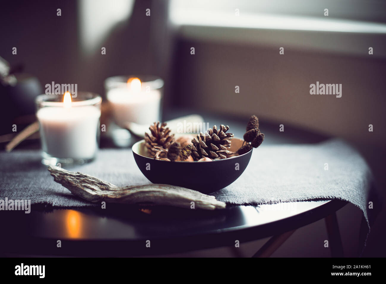 Burning candles and natural decor of cones, nuts in black bowl on the gray fabric napkin on the black table. Cozy atmosphere at home. Kinfolk Hygge Sl Stock Photo