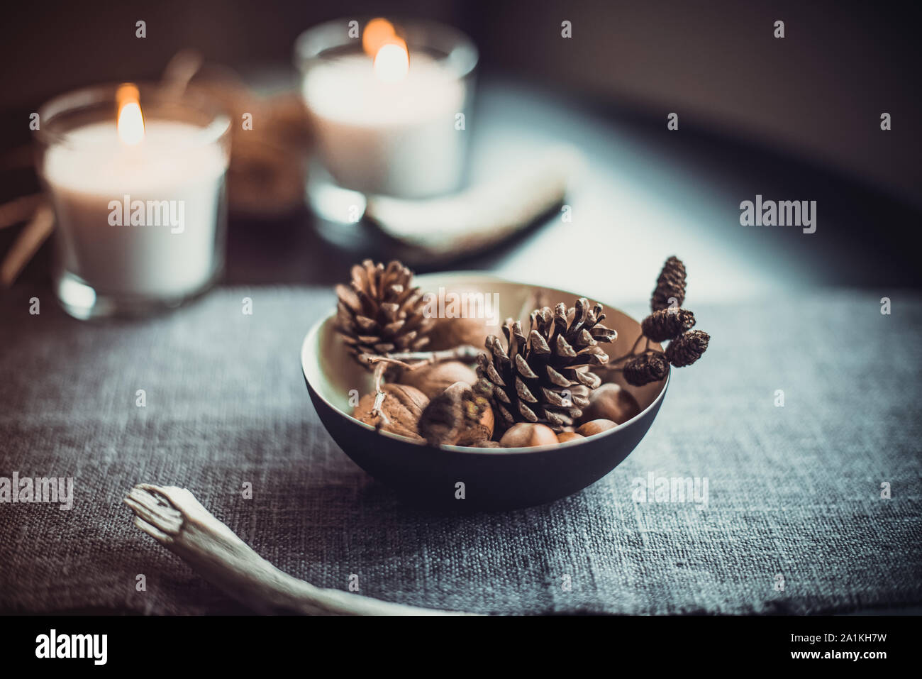 Burning candles and natural decor of cones, nuts in black bowl on the gray fabric napkin on the black table. Cozy atmosphere at home. Kinfolk Hygge Sl Stock Photo