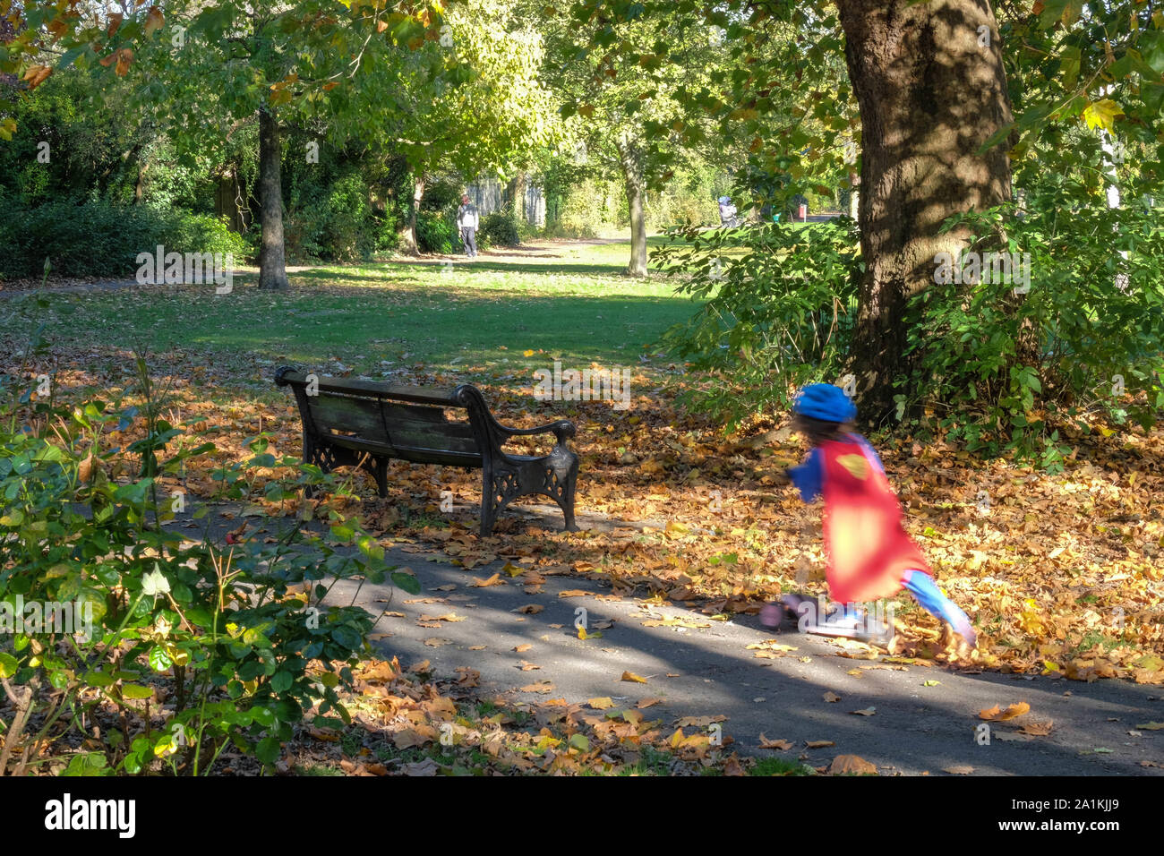 A little girl on a scooter whizzes by, dressed in a Super Hero costume at Stonegrove park in Autumn. Stock Photo