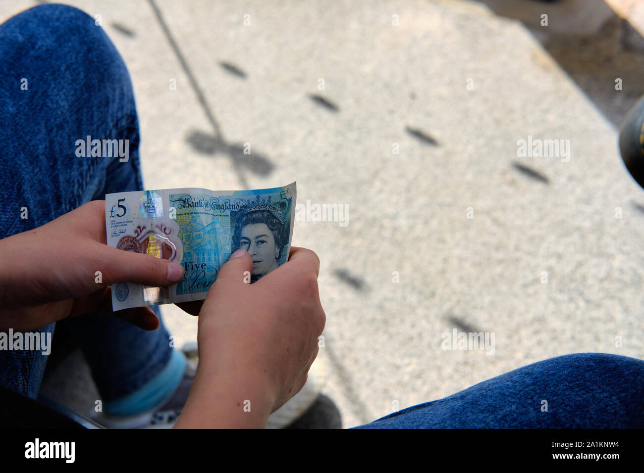 Leeds / UK - July 22nd 2019: A young person sits with a five pound sterling plastic note in their hand on a sunny day. Stock Photo