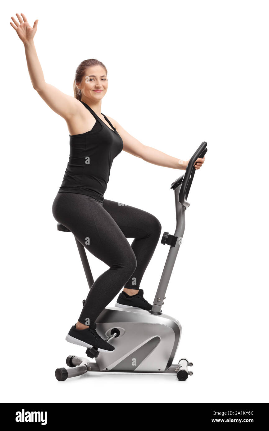 Full length profile shot of a young female riding a stationary bike and waving at camera isolated on white background Stock Photo